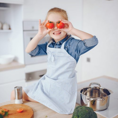 A little and beautiful girl in a blue shirt and apron is preparing a fresh vegetable salad at home in the kitchen
